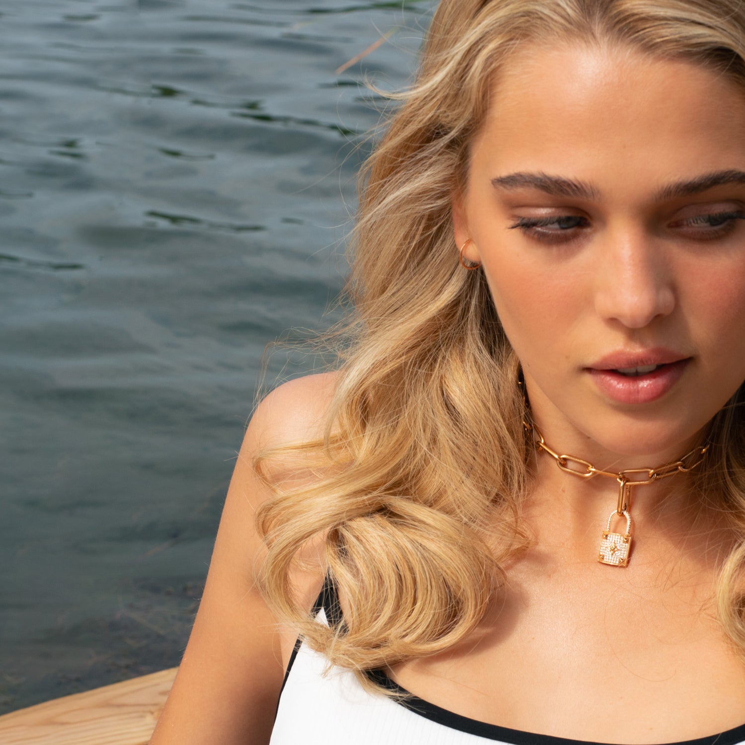 Blond woman in bikini with massive 18 KT rose gold necklace with lock pendant with white diamonds in front of blue lake