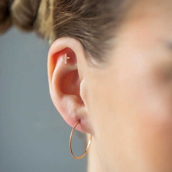 Close-up view of a woman with blond hair elegantly wearing an 18 KT rose gold cross-shaped ear stud piercing adorned with brilliant white diamonds.